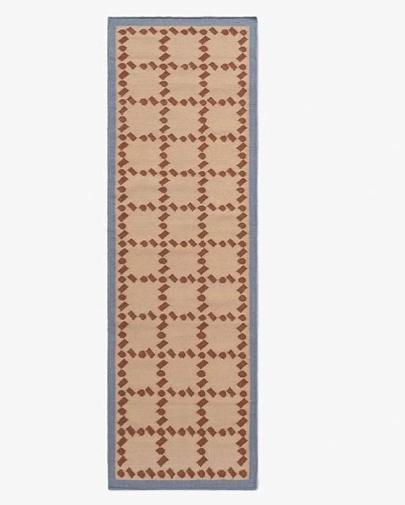 Puede incluir: Una alfombra rectangular beige con un patr&oacute;n geom&eacute;trico repetitivo en marr&oacute;n. La alfombra tiene un borde azul. El patr&oacute;n consiste en cuadrados y puntos. Esta alfombra es adecuada para un pasillo o entrada.