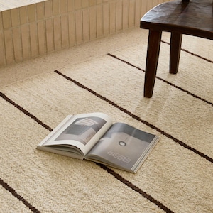 May include: A cream-colored rug with dark brown stripes. An open book rests on the rug, near a wooden chair. The rug has a textured appearance, and the stripes run horizontally across the surface.