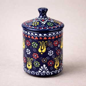 Könnte beinhalten: Ein zylindrisches Keramikgefäß mit Deckel, mit einem dunkelblauen Hintergrund, der mit bunten Blumenmustern in Gelb, Rot, Grün und Weiß verziert ist. Das Gefäß hat einen passenden Deckel mit einem kleinen Knopf und dekorativen Verzierungen.