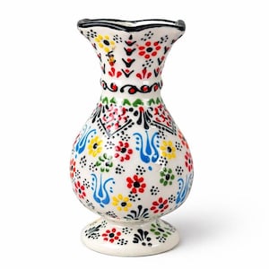 Könnte beinhalten: Eine weiße Keramikvase mit ausgestellter Oberseite und rundem Sockel. Die Vase ist mit einem farbenfrohen Blumenmuster mit roten, gelben, blauen und grünen Blumen verziert. Schwarze und rote Akzente vervollständigen das Design.