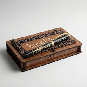Könnte beinhalten: Eine Holzbox mit dekorativer Oberseite und einem schwarz-goldenen Stift. Auf der Box ist der Name "Ismail T." in Weiß eingraviert. Der Stift hat goldene Akzente und den Namen "Yunus SEN" aufgedruckt.