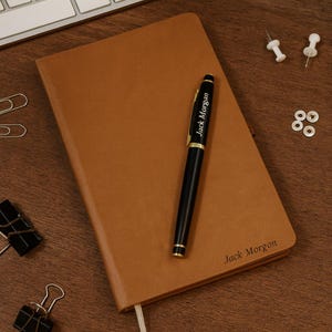 Könnte beinhalten: Ein braunes Lederjournal mit einem schwarzen Stift mit goldenen Akzenten und dem Namen "Jack Morgan". Das Journal hat auch den Namen "Jack Morgon" auf der unteren rechten Ecke. Der Stift liegt auf dem Journal.