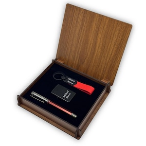 Pode incluir: Uma caixa de presente de madeira com interior preto contém uma caneta vermelha, um isqueiro preto e um chaveiro com uma alça vermelha. Cada item é gravado com o nome "Michael Brown". A caixa tem uma tampa articulada e um acabamento de grão de madeira natural.