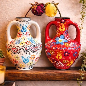 Könnte beinhalten: Zwei dekorative Keramikvasen mit Henkeln, eine weiße und eine rote, verziert mit bunten Blumenmustern. Jede Vase enthält eine getrocknete Blume. Die Vasen stehen auf einem Holzregal vor einer neutralen Wand.