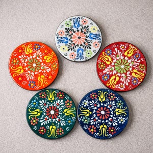 Könnte beinhalten: Fünf runde Keramikuntersetzer mit farbenfrohen Blumenmustern. Die Untersetzer weisen eine Vielzahl von Farben auf, darunter Rot, Orange, Grün, Blau und Weiß. Jeder Untersetzer hat ein einzigartiges Muster mit einem zentralen Blumenmotiv.