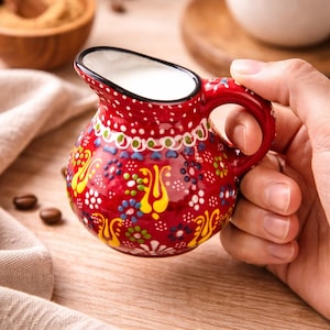 Könnte beinhalten: Eine kleine, rote Keramik-Sahnekännchen mit Henkel und Ausguss, gefüllt mit Milch. Das Sahnekännchen ist mit einem bunten Blumenmuster verziert. Der Hintergrund umfasst eine Tasse Kaffee, Kaffeebohnen und eine Holzschale.