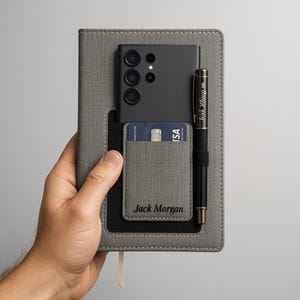 Könnte beinhalten: Ein graues Notizbuch mit Telefon, Stift und Kartenhalter. Das schwarze Telefon und der Stift sind sichtbar. Eine Kreditkarte befindet sich im Kartenhalter, und der Name "Jack Morgan" ist auf dem Halter aufgedruckt.