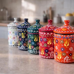 Könnte beinhalten: Fünf Keramikbehälter mit Deckeln, verziert mit bunten Blumenmustern. Die Behälter sind in verschiedenen Farben erhältlich: Weiß, Marineblau, Grün, Rot und Orange. Jeder Deckel hat einen kleinen Knopf.