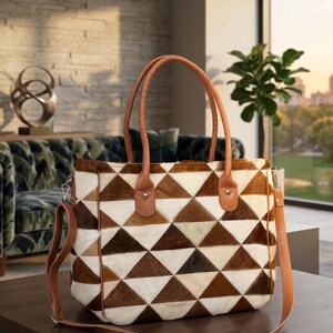 May include: A tote bag featuring a geometric pattern of brown, tan, and white triangular hair-on-hide patches. The bag includes two tan leather top handles, a detachable tan leather shoulder strap, and silver-toned metal hardware attachments.