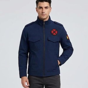 Op de afbeelding: Een marineblauw utility-jack met een hoge kraag, een ritssluiting over de hele lengte en twee borstzakken met kleppen. Op de linkerborst zit een ronde rode patch met een zwarte X en op de linkermouw een kleine rechthoekige patch.