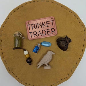 May include: A selection of fantasy-themed trinkets, including a sign reading "TRINKET TRADER", a small glass bottle, a blue potion bottle, a raven figurine, a blue gem, a small brown pouch, and a beaded charm, arranged on a tan leather-like surface.