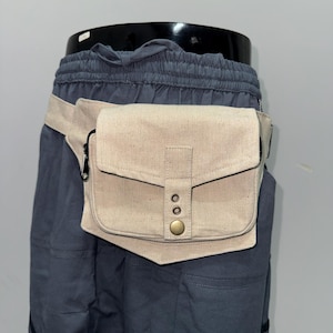 May include: A beige canvas fanny pack with a flap closure and a brass button. The bag is worn around the waist with a matching beige strap. The fanny pack is attached to a pair of dark blue shorts.