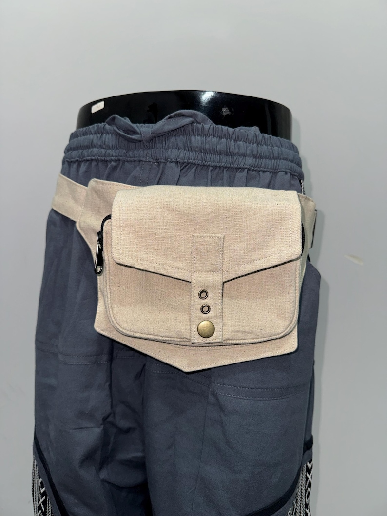 May include: A beige canvas fanny pack with a flap closure and a brass button. The bag is worn around the waist with a matching beige strap. The fanny pack is attached to a pair of dark blue shorts.