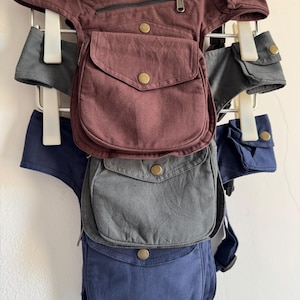 May include: Three canvas utility hip bags in burgundy, charcoal grey, and navy blue. Each bag features a main flap pocket with a brass-tone snap button, a zippered compartment, and an adjustable strap with a side-release buckle for secure attachment.