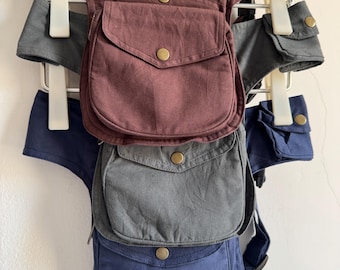 May include: Three canvas utility hip bags in burgundy, charcoal gray, and navy blue. Each bag features a main flap pocket with a brass-tone snap button, a zippered compartment, and an adjustable strap with a side-release buckle for secure attachment.