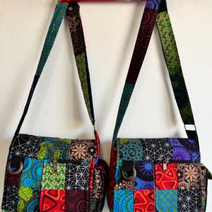 May include: Two patchwork messenger bags with patterned straps hang against a white wall. The bags feature a variety of colours and patterns, including blue, green, red, and black. Each bag has a front pocket with a zip and a metal ring.