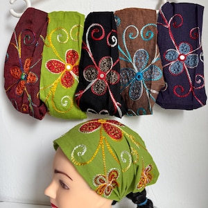 Op de afbeelding: Geborduurde haarbanden in diverse kleuren, waaronder bordeauxrood, groen, zwart, bruin en paars, hangend aan een witte hanger. Elke haarband heeft een bloemenpatroon met contrasterende draden. Een mannequin draagt een groene haarband.