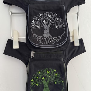 Puede incluir: Dos riñoneras negras con correas ajustables y compartimentos con cremallera. La bolsa superior presenta un diseño de árbol de la vida en blanco con hojas en forma de corazón, mientras que la inferior muestra un diseño en color verde.