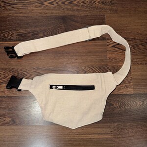 May include: Beige fanny pack with a black zipper and adjustable strap. The fanny pack has a black buckle closure and is made of a woven fabric. The strap is approximately 40 inches long.