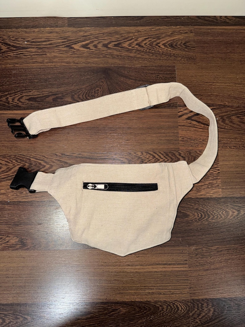 May include: Beige fanny pack with a black zipper and adjustable strap. The fanny pack has a black buckle closure and is made of a woven fabric. The strap is approximately 40 inches long.