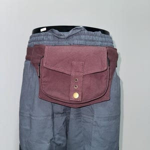 May include: A burgundy fanny pack with a rectangular pocket and a flap secured by a brass button. The pack has a zippered pocket and is attached to a burgundy belt. The pack is worn over gray pants.
