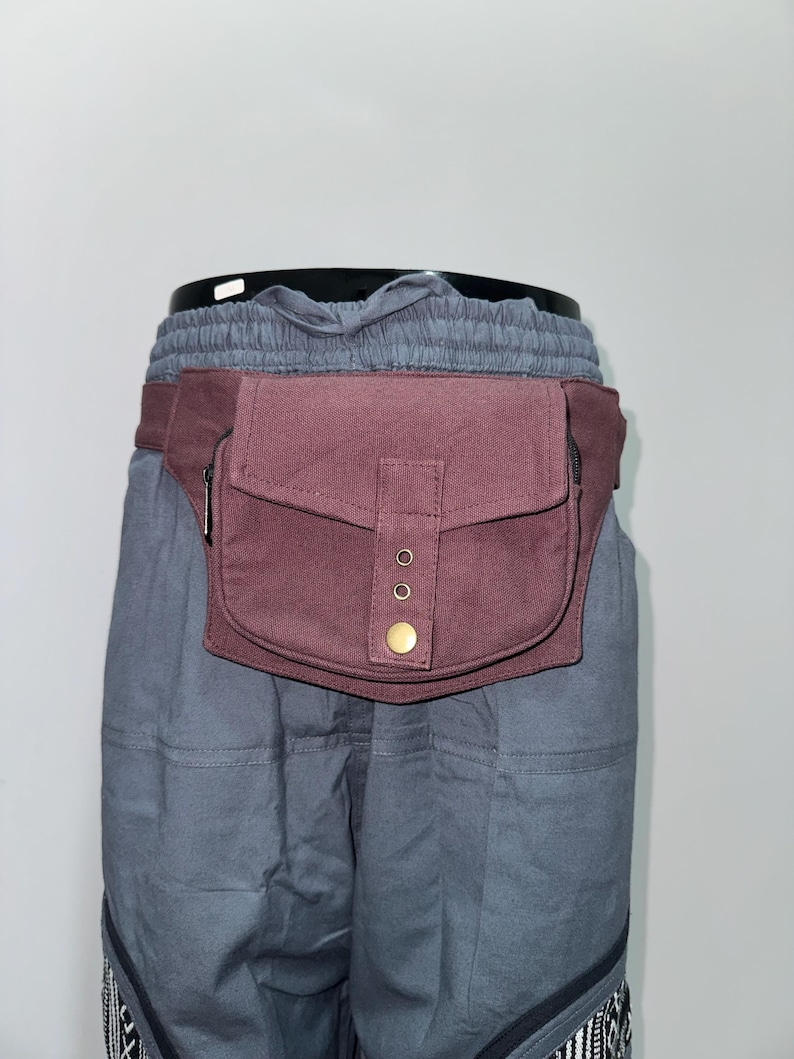 May include: A burgundy fanny pack with a rectangular pocket and a flap secured by a brass button. The pack has a zippered pocket and is attached to a burgundy belt. The pack is worn over gray pants.