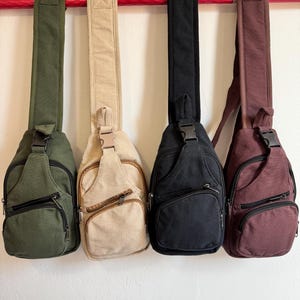 May include: Four canvas sling bags in olive green, beige, black, and burgundy hang from a red bar. Each bag has a zippered front pocket, adjustable strap, and a buckle closure. The bags are designed to be worn across the body.
