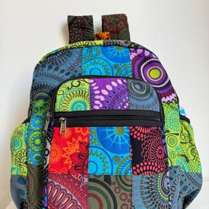 May include: A colorful patchwork backpack with a variety of patterns and colors, including blue, green, red, and purple. The backpack has a zippered front pocket, side pockets, and adjustable straps. The backpack is made of fabric.