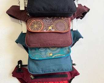 May include: A display of several fanny packs in various colors, including black, burgundy, teal, and red. Each pack features a zippered main compartment and a front pocket with decorative embroidery or patterns. The packs are hanging on a white rack.