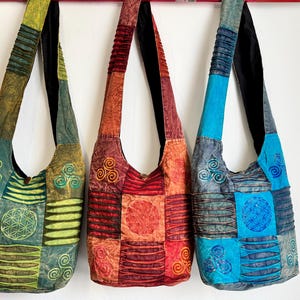 Peut inclure: Trois sacs à bandoulière de style hobo suspendus en rangée, chacun présentant un design en patchwork avec des panneaux de tissu texturés. Les sacs sont colorés dans des tons de vert, rouge orangé et bleu, ornés de motifs triskèles et géométriques brodés.