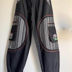 Könnte beinhalten: Schwarze Hose mit elastischem Kordelzugbund und Bündchen an den Knöcheln. Die Hose verfügt über große seitliche Cargotaschen mit brauner Umrandung, die mit gestickten Pilzmotiven und schwarz-weiß gemusterten Stoffeinsätzen verziert sind.