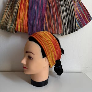 Op de afbeelding: Een collectie van zeven brede, geribbelde stoffen hoofdbanden in diverse meerkleurige patronen, waaronder oranje, gele, paarse en zwart-witte strepen, gepresenteerd op een wit etalagehoofd en hangend aan een witte plastic kledinghanger.