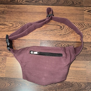 May include: A burgundy fanny pack with a black zipper and adjustable strap. The pack is made of a soft fabric and has a black plastic buckle. The fanny pack is designed to be worn around the waist or across the body.