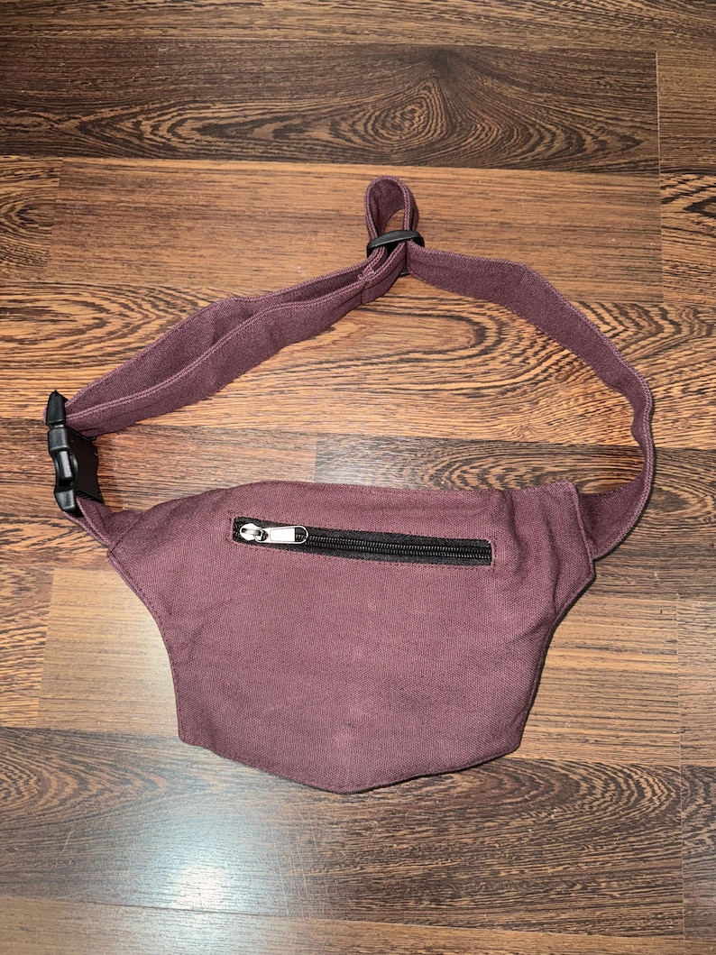 May include: A burgundy fanny pack with a black zipper and adjustable strap. The pack is made of a soft fabric and has a black plastic buckle. The fanny pack is designed to be worn around the waist or across the body.
