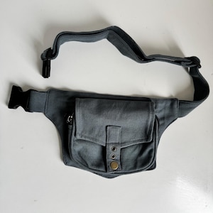 Könnte beinhalten: Eine graue Canvas-Bauchtasche mit einem schwarzen, verstellbaren Riemen und einer Schnalle. Die Tasche hat eine Vordertasche mit einer Klappe, die mit zwei Druckknöpfen und einem Messingknopf befestigt ist. Ein kleiner Reißverschluss ist an der Seite der Vordertasche sichtbar.
