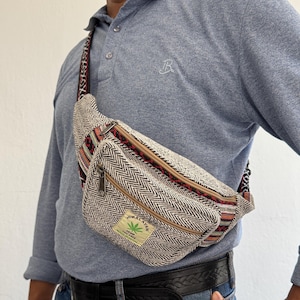 May include: A beige and black herringbone patterned bum bag with a front zippered pocket and a woven strap. The bag features a rectangular patch with the text HIMALAYAN, a green leaf icon, and the words HEMP THC FREE printed in black ink.