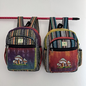 Puede incluir: Dos mochilas con patrones de rayas verticales en negro, blanco y varios colores. Cada bolsa tiene un bolsillo frontal con efecto tie-dye y un diseño de setas bordado. Una pequeña etiqueta en el bolsillo dice HIMALAYAN HEMP THE FREE.