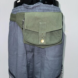 May include: An olive green fanny pack with a zippered pocket and a flap closure secured by a brass button. The pack is attached to a matching green belt. The fanny pack is worn over gray pants with a patterned design at the bottom.