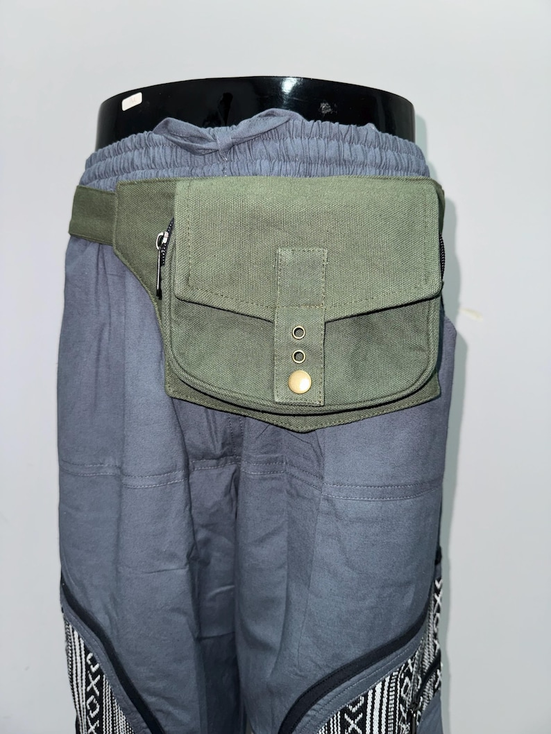 May include: An olive green fanny pack with a zippered pocket and a flap closure secured by a brass button. The pack is attached to a matching green belt. The fanny pack is worn over gray pants with a patterned design at the bottom.
