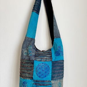 Può includere: Una borsa a tracolla in stile hobo con un design patchwork nei toni del blu e del grigio. La borsa presenta simboli triskelion ricamati e un motivo centrale a fiore della vita, accentuato da pannelli di tessuto plissettato e una lunga tracolla.