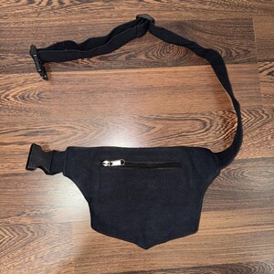 May include: Black fanny pack with a zippered pocket and adjustable strap. The pack has a curved shape and a buckle closure. The strap is made of a durable, black fabric. The fanny pack is designed to be worn around the waist.
