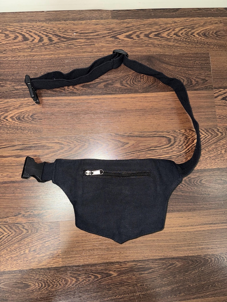 May include: Black fanny pack with a zippered pocket and adjustable strap. The pack has a curved shape and a buckle closure. The strap is made of a durable, black fabric. The fanny pack is designed to be worn around the waist.
