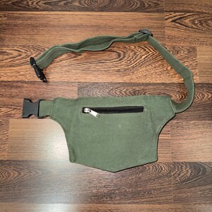 May include: An olive green fanny pack with a black zipper and adjustable strap. The pack has a rectangular shape with a curved bottom and a black plastic buckle closure. The strap is made of the same olive green material.