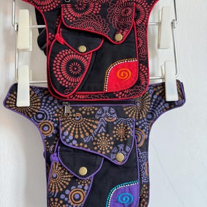 Pode incluir: Duas bolsas de perna com múltiplos bolsos com fechos de pressão e compartimentos com zíper. A bolsa superior é preta com padrões florais vermelhos e um detalhe vermelho. A bolsa inferior é preta com padrões roxos e marrons.