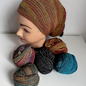 Può includere: Un cappello lavorato a maglia multicolore è esposto su una testa di manichino, con diversi gomitoli di filato abbinati in vari colori. Il cappello e i gomitoli presentano un mix di tonalità rosse, gialle e marroni, con un po' di verde acqua e nero.