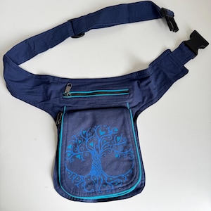 Pode incluir: Cinto de cintura azul marinho com um desenho de árvore da vida. Possui um compartimento principal com zíper e um bolso frontal com acabamento turquesa. A alça ajustável tem uma fivela de plástico preta. O desenho da árvore é em tons de azul.