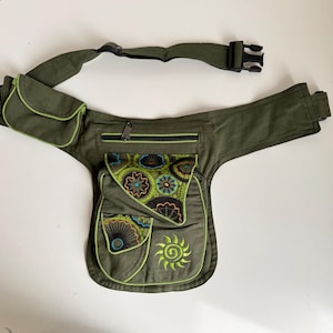 May include: An olive green waist bag with multiple pockets and a patterned flap. The bag features a zipped main compartment, two smaller pockets, and a decorative flap with a floral design. It has a black buckle closure and adjustable strap.