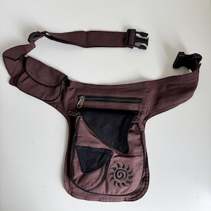 Pode incluir: Cinto de cintura bordô com vários bolsos e fecho de fivela preto. A bolsa possui um bolso com zíper preto e um desenho de sol preto bordado na frente. A alça ajustável permite um ajuste confortável.