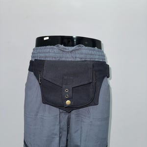 May include: Black fanny pack with a flap closure and brass-colored rivets. The pack is worn around the waist and is attached to gray pants. The pants have a drawstring waist and decorative patterned trim at the bottom.