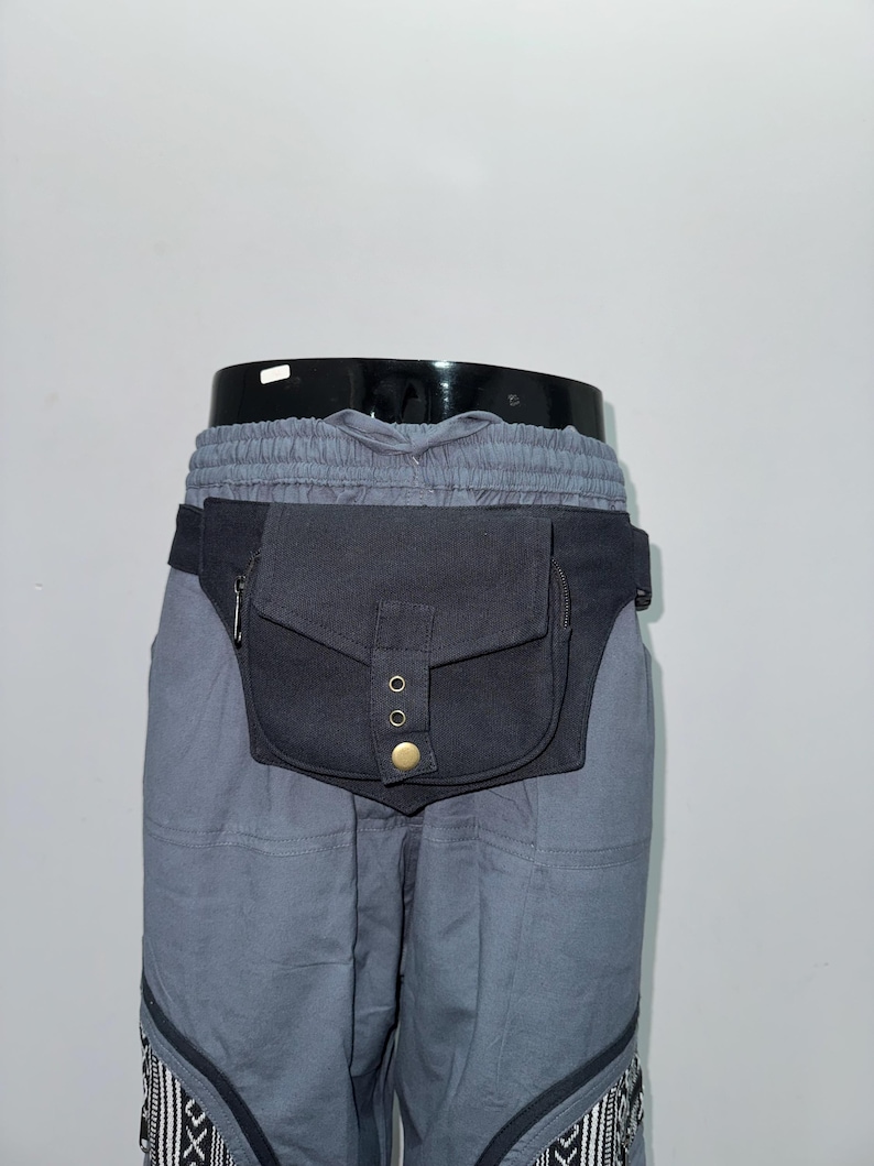 May include: Black fanny pack with a flap closure and brass-colored rivets. The pack is worn around the waist and is attached to gray pants. The pants have a drawstring waist and decorative patterned trim at the bottom.